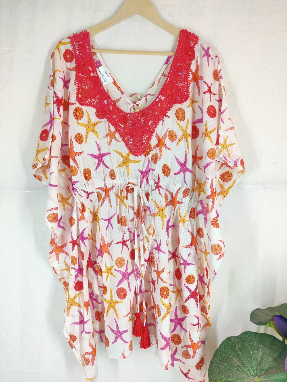 White cotton kaftan printed with a pink, orange and yellow starfish and shell motif, with a red crochet lace yoke at the neckline and red tassels on the waist tie
